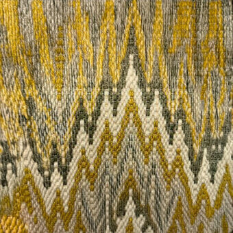 Geometric textile with zigzag pattern in olive green, creamy white, and mustard yellow; dense weave with visible, textured threads.