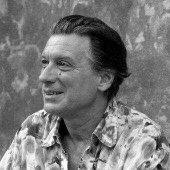 Black and white portrait of Ferdi Giardini, a man with graying hair, wearing a floral patterned shirt, against a textured gray wall.
