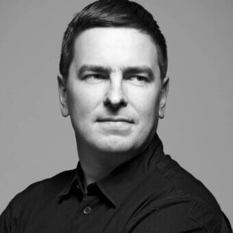 Black and white portrait of Harri Koskinen, a man with short dark hair wearing an unbuttoned black shirt, against a graduated gray backdrop.