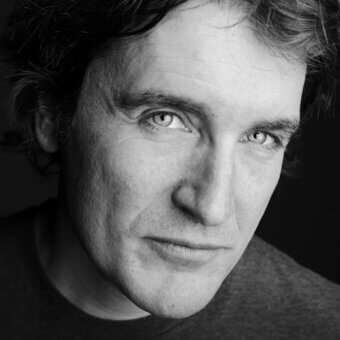 Black and white portrait of a man with tousled dark hair, wearing a dark gray knit garment, contemplative gaze, blurred inscription.