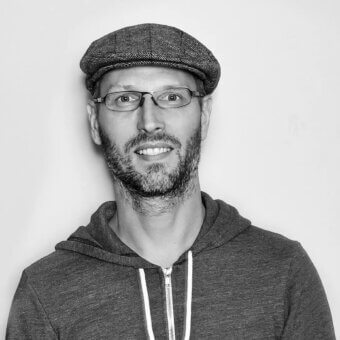 Black and white portrait of Lutz Pankow wearing a gray tweed cap and glasses, with a dark gray hooded sweatshirt, against a white backdrop.