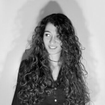 Black and white portrait of a woman with long, dark curly hair, wearing a v-neck shirt and choker necklace, gentle smile, studio lighting.