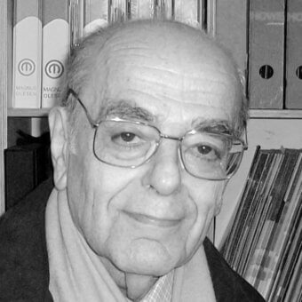 Portrait of an elderly man in glasses and scarf, with stacked file folders labeled "Magnus Olesen" in a neutral office setting.