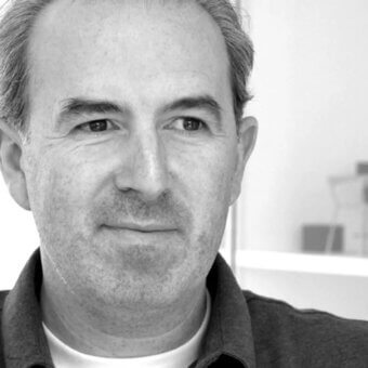Black and white portrait of Sam Hecht, a man with graying hair wearing a white t-shirt and dark textured jacket, neutral expression.