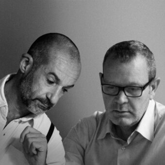 Black and white portrait of two mature men in white shirts against a light gray backdrop, one gesturing with hand on dark strap.