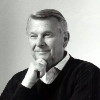Black and white portrait of Tito Agnoli, a man with gray hair wearing a dark sweater and striped collared shirt, hand resting on chin.