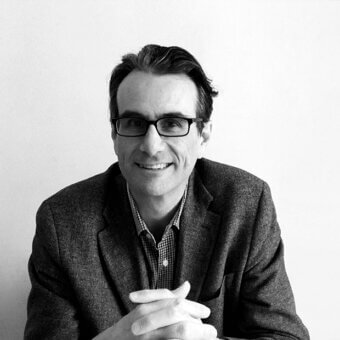 Carlo Tamborini portrait, man in gray tweed blazer and checkered shirt, wearing black glasses, against a light neutral wall.