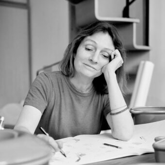 Black and white portrait of Inga Sempé at a desk with sketches, wearing a gray t-shirt and silver bangles, soft diffused lighting.