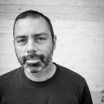 Black and white portrait of Marc Krusin with short dark hair, beard, and neutral expression against a textured white brick wall.