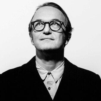Black and white portrait of Massimiliano Locatelli, a man in a blazer and glasses looking up with a thoughtful expression against a white background.