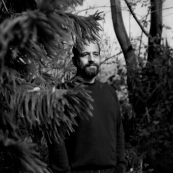 Black and white portrait of Michael Anastassiades in a dark sweater, framed by evergreen foliage, with a serious expression and diffused natural lighting.
