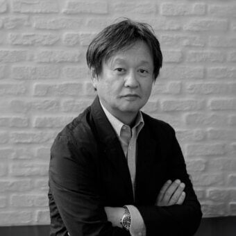 Naoto Fukasawa portrait, black and white photograph, olive shirt, navy blazer, exposed brick wall, neutral expression, soft lighting.
