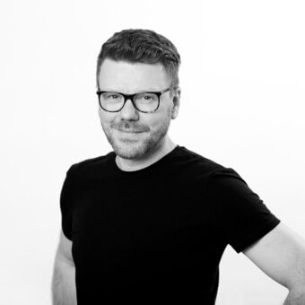 Oliver Schick portrait, black and white photograph, angled pose, short dark hair, glasses, black t-shirt, neutral expression, white background.