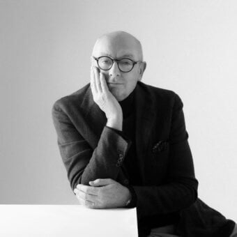 Piero Lissoni portrait, grayscale photograph, seated man in tweed blazer and turtleneck, contemplative expression, soft lighting.