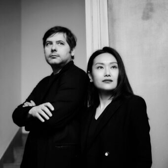 Black and white portrait of two partners in black blazers, standing before a distressed plaster wall with a visible staircase.