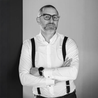 Black and white portrait of Andrea Citterio, a man with gray beard wearing white shirt, dark suspenders, and framed glasses, arms crossed.