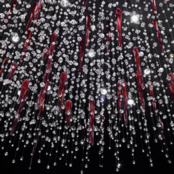 Crystal suspension lamp by Cassoni, red cylindrical accents, layered strands descend from ceiling, black background, reflective sparkle.