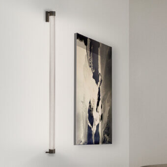 Trama wall lamp by Cassoni, dark bronze metal frame with clear glass tubes, mounted on light gray wall with abstract gray painting.