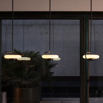 Tratto suspension lamps, bronze metal & frosted glass, elongated oval shades hang from dark rods with green accents against concrete.
