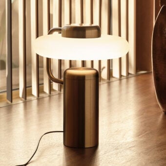 Kundalini Tratto table lamp, brushed brass finish, curved arm with white opaque shade on dark wood surface against brass slat wall.