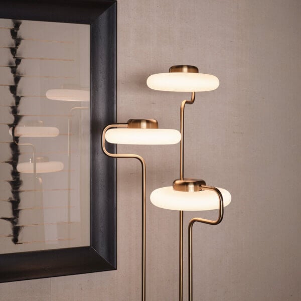 Floor lamp, brass and opal glass, three-tiered design, positioned beside framed abstract art with black and gold lines on beige textured wall.