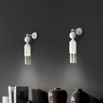 Masiero Nappe wall lamps with white shades and gold accents cast shadows on a dark gray wall, alongside books and gray ceramic vases.