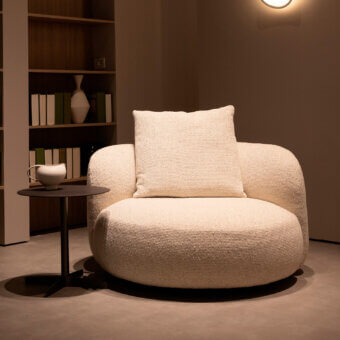 Vic lounge chair, off-white fabric, rounded cocoon shape with square cushion, dark gray base, side table, bookcase backdrop, gray interior.
