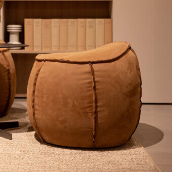 Leather ottoman, tan-brown, rounded shape with vertical seams, centered on beige textured rug with a bookshelf backdrop.