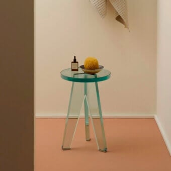 Glas Italia side table, clear glass, circular turquoise rim, tripod legs, amber bottle, beige sponge, terracotta floor, beige wall.