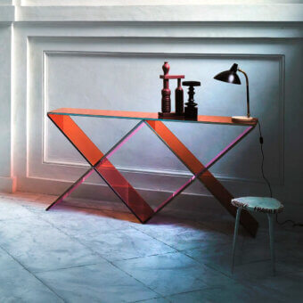 Glas Italia XX console table, orange glass top, geometric reddish-pink base, studio setting, desk lamp, stool with “FRAGILE” text.
