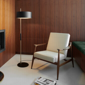 Cassoni Oluce floor lamp, gold stem, black shade, against dark wood paneling, with mid-century armchair and magazine stack.