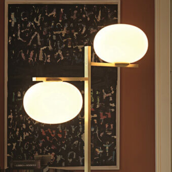 Oluce Alba floor lamp, brushed gold finish, with two matte white spherical shades, against a terracotta wall and layered paper collage.