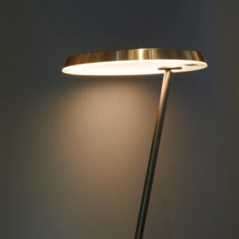 Amanita floor lamp, brushed metal, with a large circular lampshade and brass rim, emitting warm diffused light against a dark gray wall.