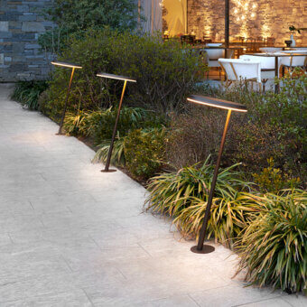Amanita suspension lamps in dark gray metal illuminate a stone pathway bordered by greenery, with a dining area visible in the background.