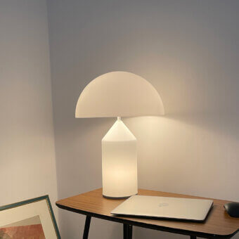 Atollo glass table lamp with frosted glass shade and cylindrical base on light wood table, silver laptop nearby.