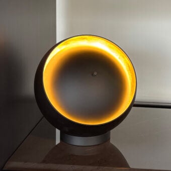Eva table lamp, dark gray metal, spherical shape with amber light; polished marble surface and slender metallic stem visible.