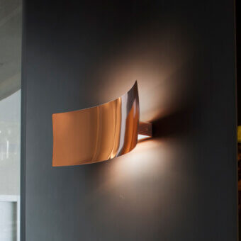 Oluce Lens Wall Lamp, copper finish, curved shade emits warm light on dark gray wall, minimalist interior setting, brushed metallic texture.