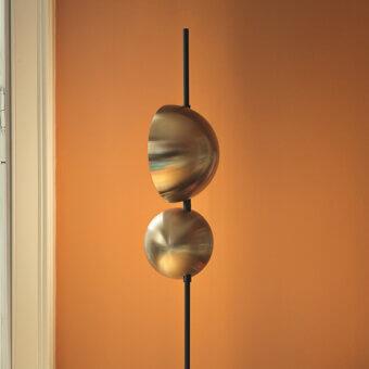 Cassoni Oluce Superluna floor lamp, brushed brass, two vertically stacked semi-spherical shades on black metal rod against orange wall.