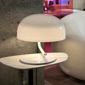 Cassoni Oluce table lamp, white dome shade, marble base with gray veining, slender white arm, against textured concrete wall.