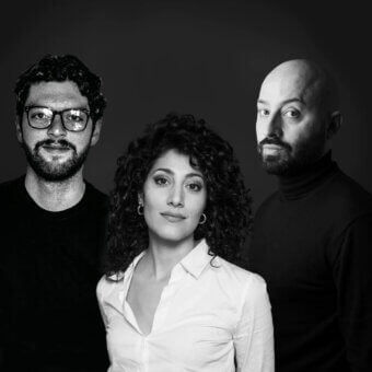 Black and white portrait of three people—man, woman, and man—with curly hair, glasses, and beards against a dark gray backdrop.