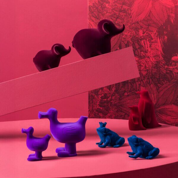 Animal figurines—magenta elephants, purple ducklings, blue frogs, red penguin—arranged on red geometric platforms against a red botanical print backdrop.