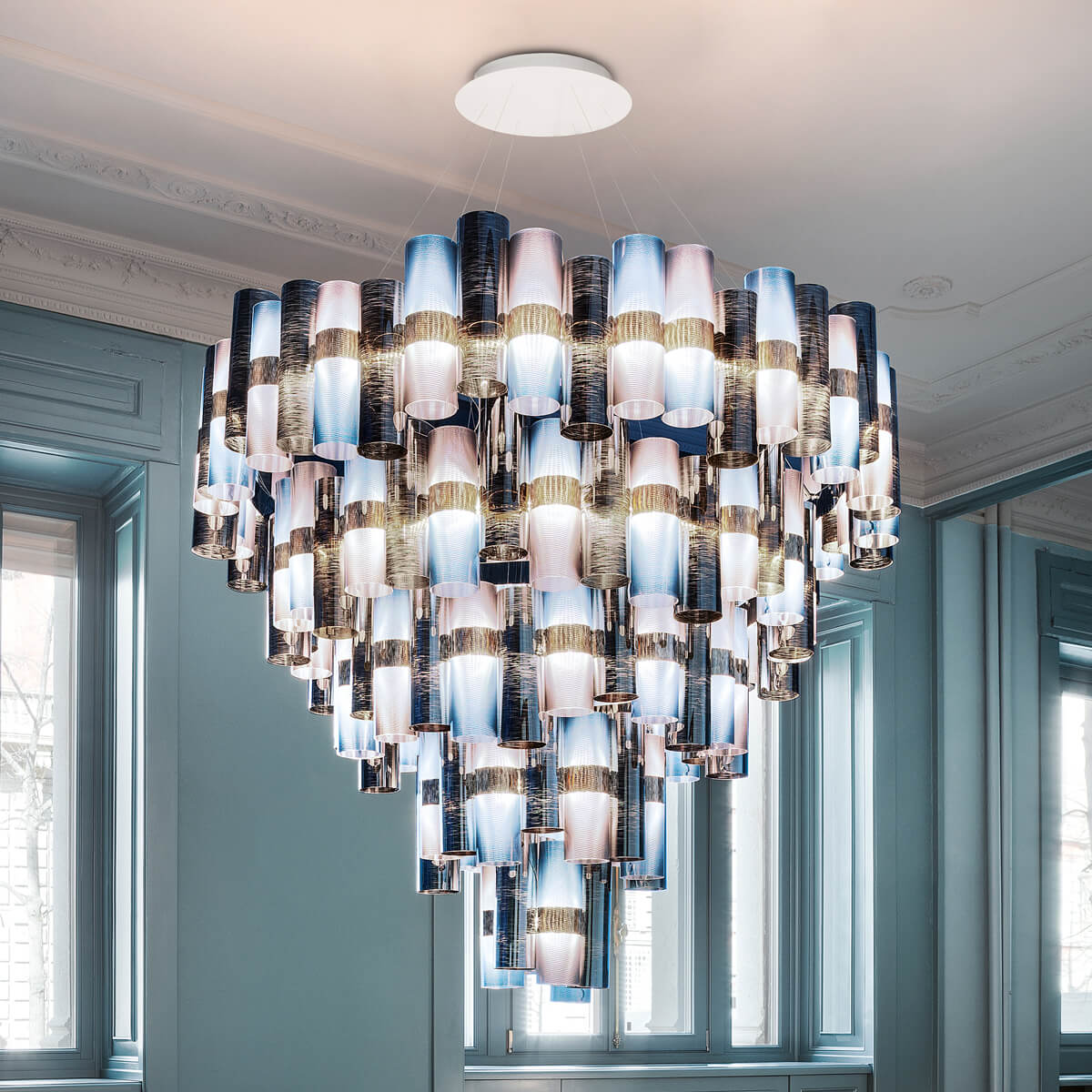 Chandelier, iridescent fabric, tiered cylindrical shades cascade from a white ceiling mount in a room with pale blue walls and green window frames.