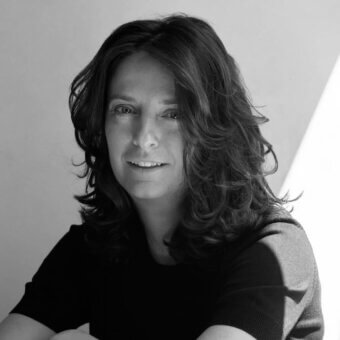 Black and white portrait of a woman with wavy dark hair, wearing a black crew-neck sweater, against a light wall.