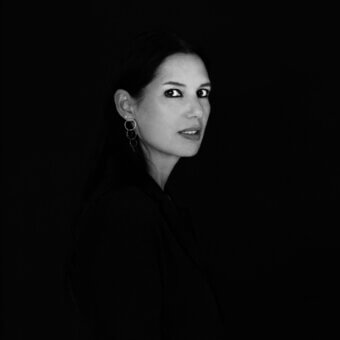 Black and white portrait of Elisa Ossino, three-quarter profile, dark collared shirt, silver hoop earrings, minimalist composition, diffused lighting.
