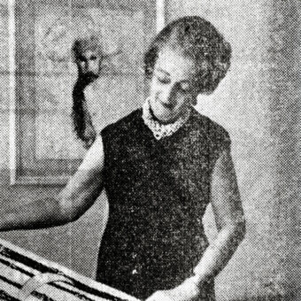 Woman behind table with rectangular objects, wearing dark dress and beaded necklace, gesturing in diffused light interior.