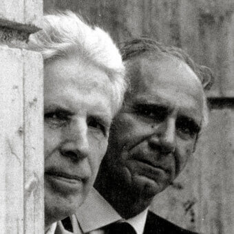 Black and white portrait of two men in suits, framed by a weathered wood post, with high contrast and blurred outdoor background.