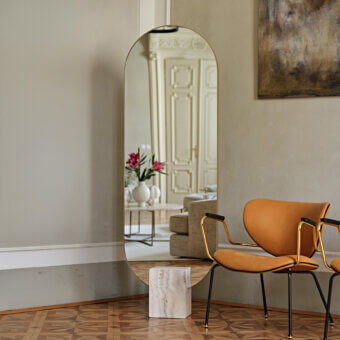 Captivating Tìam mirror crafted in Italy with a gracefully curved golden metallic frame and majestic travertine base