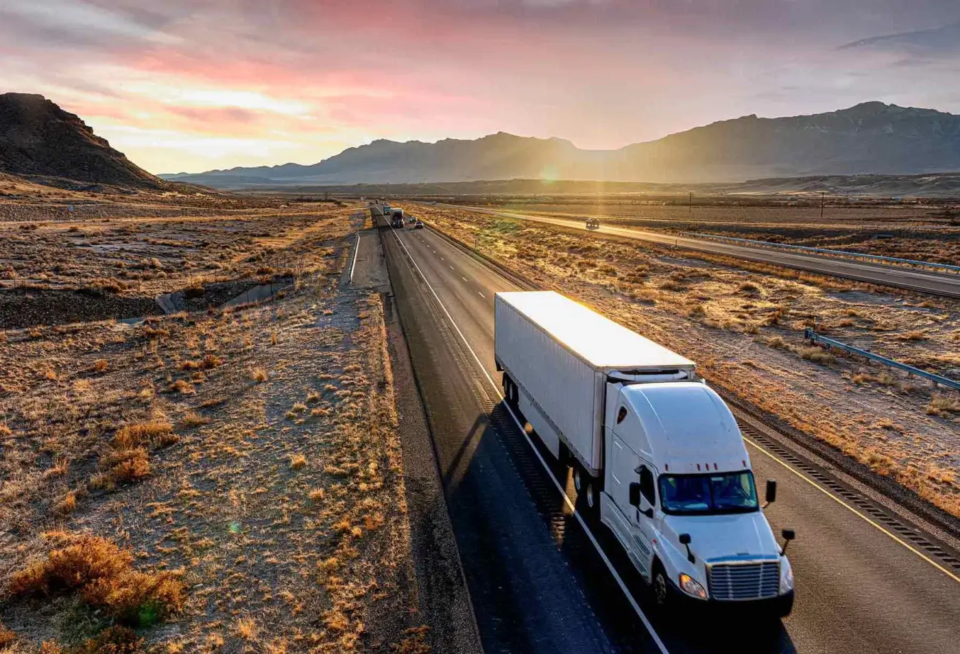 Desert highway scene with a white semi-truck traveling towards a pink, orange, and yellow sunrise over arid terrain and distant mountains.