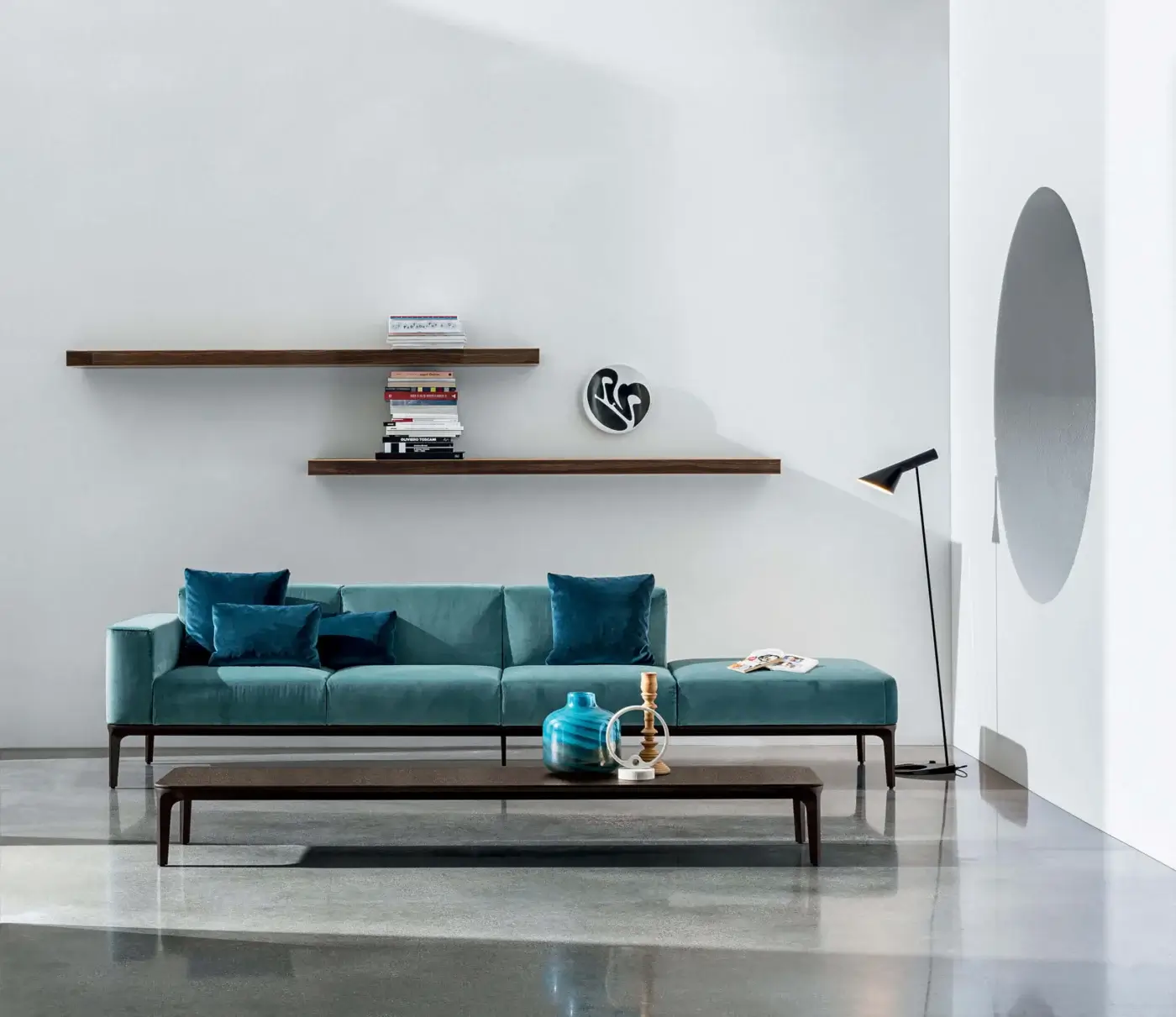 Teal sofa with curved back and metal frame, paired with a rectangular table and dark wood shelf displaying books and floral art.