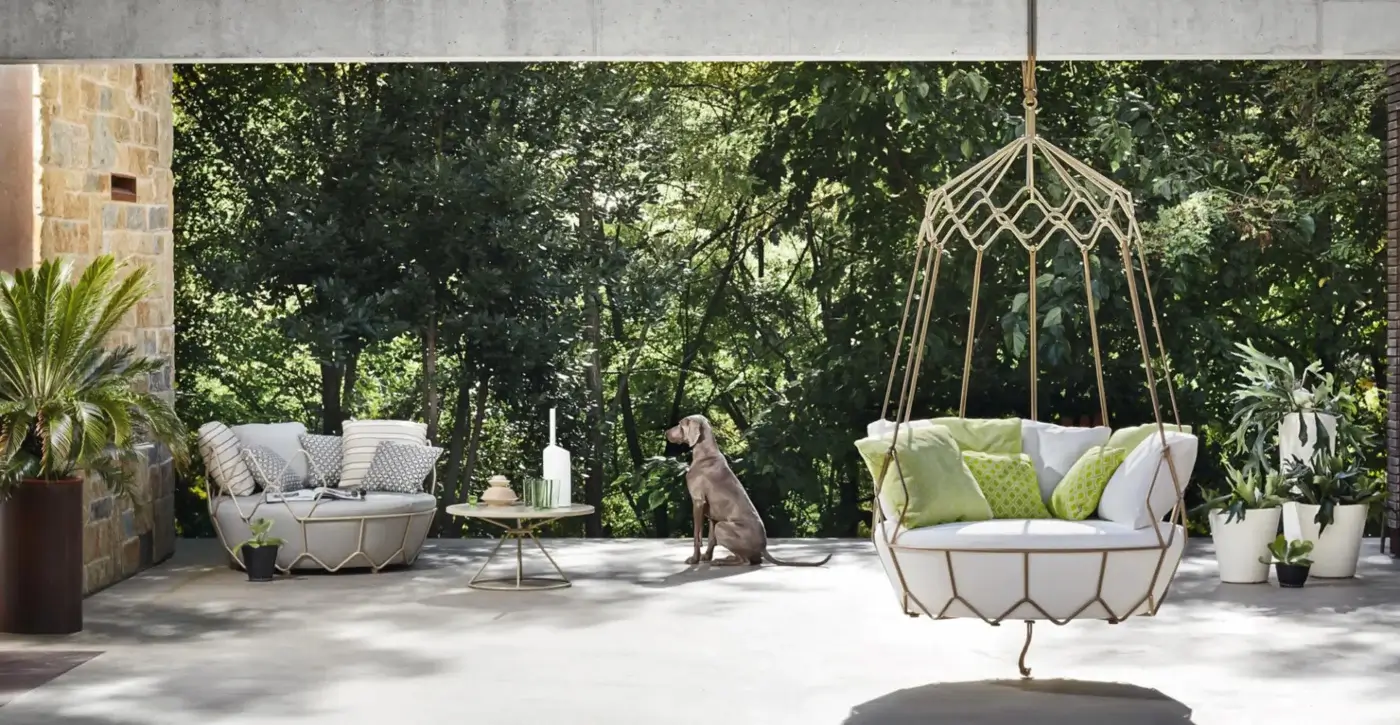 Rattan lounge chair suspended from gold metal frame, white woven exterior, light-green cushions, concrete patio, green foliage backdrop.